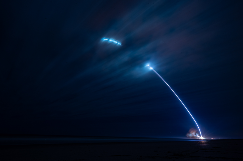 A long-exposure night photograph traces a rocket's ascent as a bright white arc climbing away from a coastal launch pad, with a second detached arc burning higher in the sky.