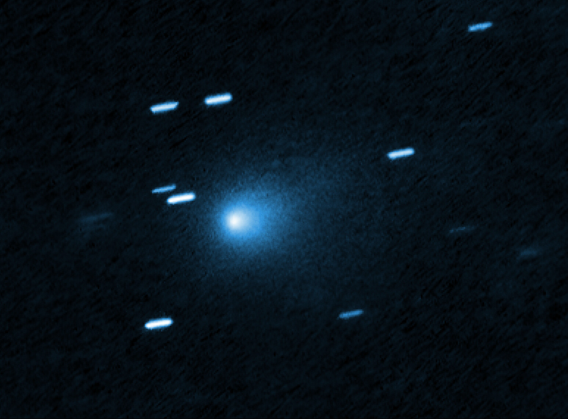 A telescope exposure shows a comet as a compact bright core inside a soft blue coma, while background stars are drawn into short dashes by the tracking.