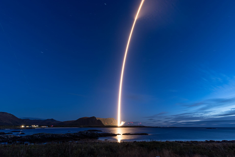 A bright rocket exhaust trail curves upward from a launch site beside a dark, calm sea and mountainous shoreline under a blue evening sky.