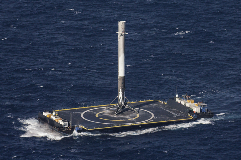 A Falcon 9 first stage stands upright on the deck of an autonomous drone ship in open ocean, seen from the air with tugs alongside.