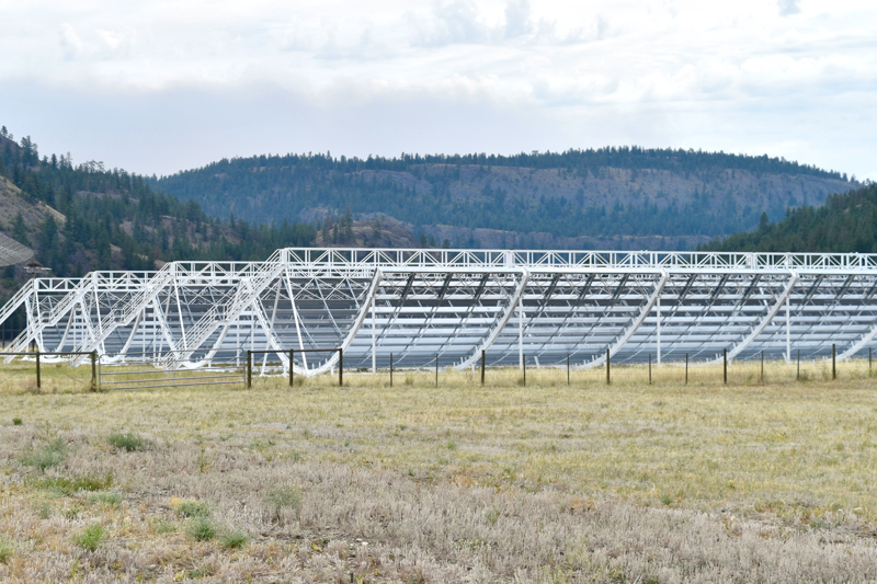 The CHIME telescope's four 100-metre wire-mesh cylindrical reflectors in an open field near Penticton, British Columbia, with forested hills behind. Image: Wikimedia Commons, CC BY-SA 4.0.