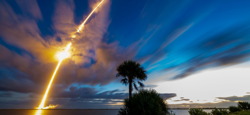 A long-exposure photograph of a SpaceX Falcon 9 climbing through the pre-dawn sky from Cape Canaveral, the exhaust plume forming a bright diagonal streak through scattered cloud above silhouetted palm trees. Source: SpaceX, via the Bandwagon-5 mission page. Full-size file: https://sxcontent9668.azureedge.us/cms-assets/assets/20261020_CC_Band_5_DSC_0759_desktop_81035d0e4c.jpg