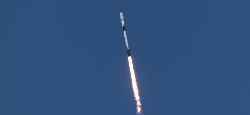 Falcon 9 ascends against a deep blue sky over Vandenberg, the bright plume trailing straight down from the booster as it climbs out of the lower stage of the atmosphere.