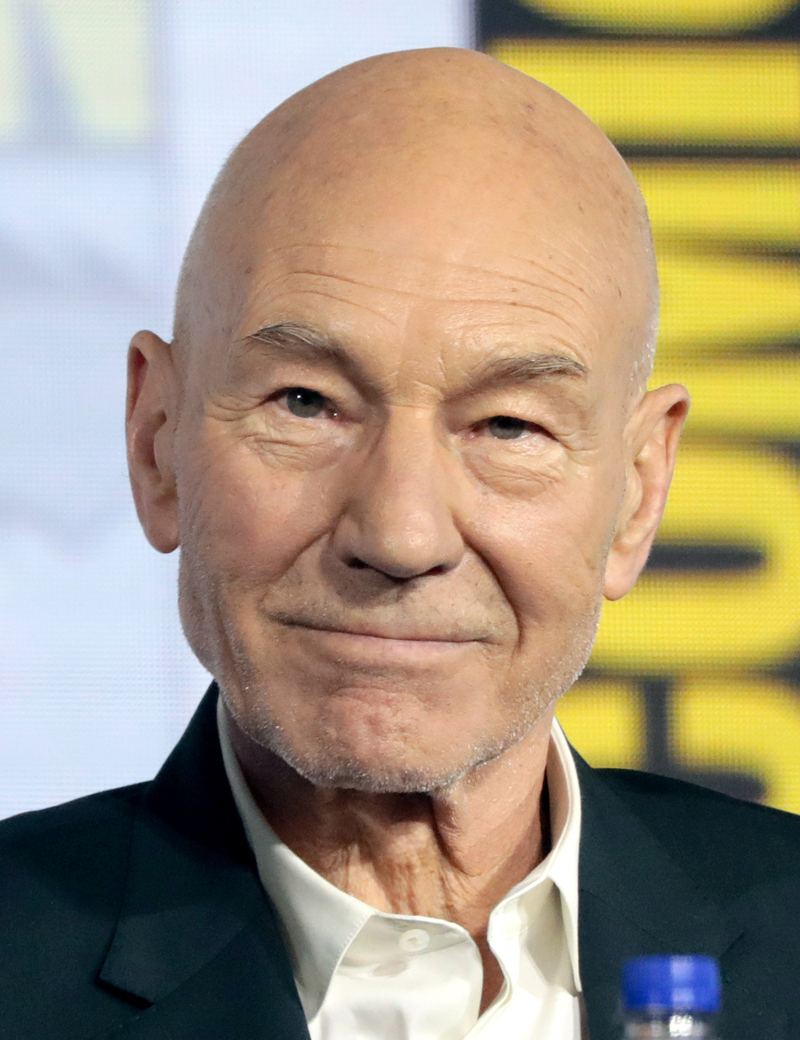 Patrick Stewart speaking at the 2019 San Diego Comic-Con International, wearing a dark jacket over an open-collared white shirt with a yellow and black backdrop behind him (Gage Skidmore, CC BY-SA 3.0, via Wikimedia Commons)